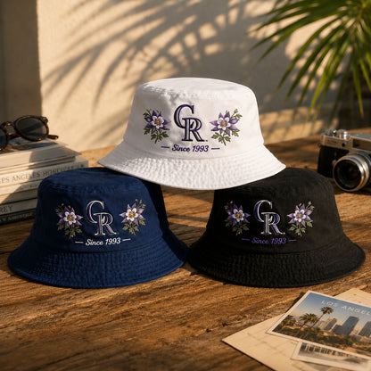 Three embroidered bucket hats in white, navy blue, and black, arranged on a rustic wooden table with a vintage camera, sunglasses, and books in the background.